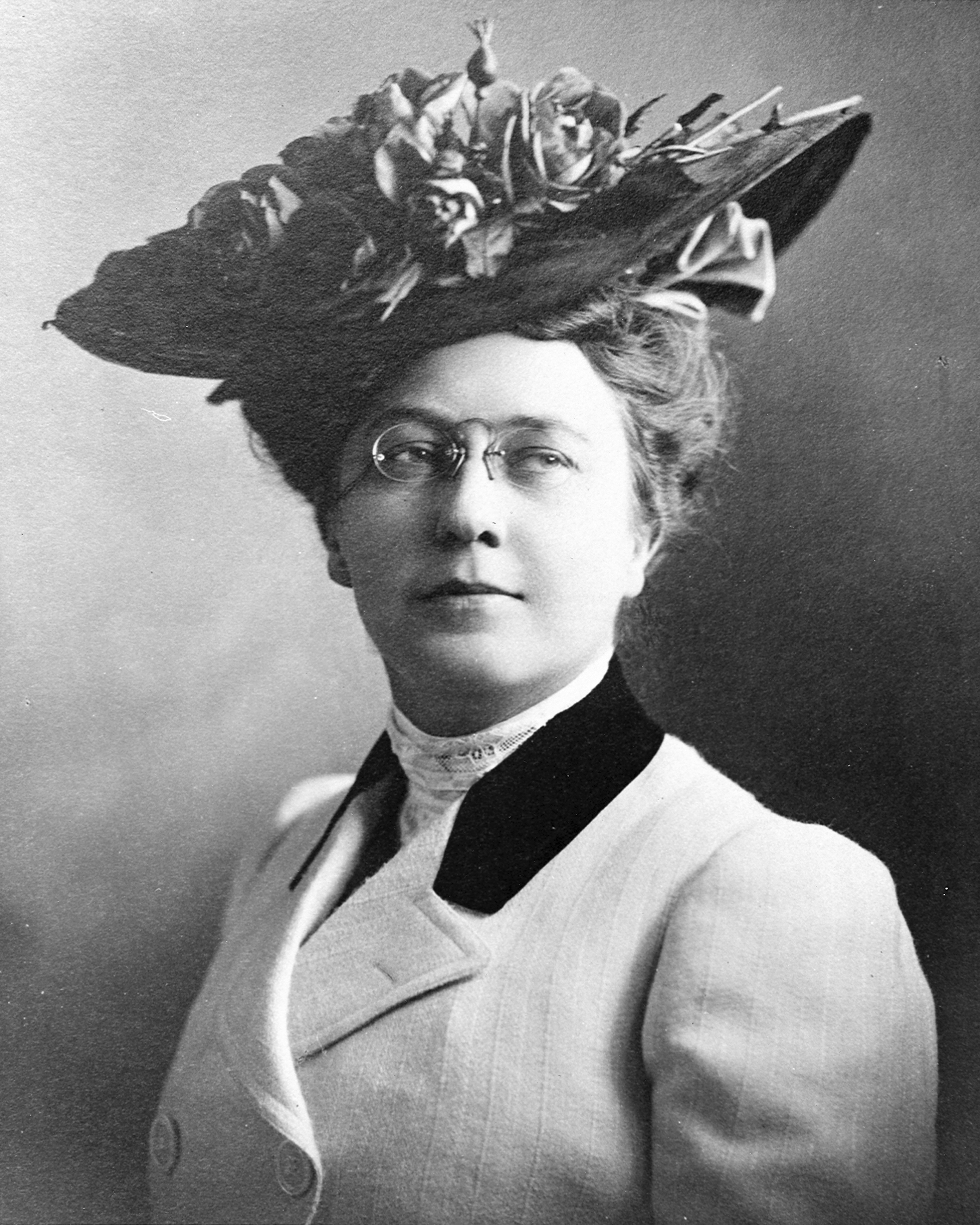 Woman facing camera, close-up portrait in black and white, with a large hat and glasses, wearing a white double breasted coat